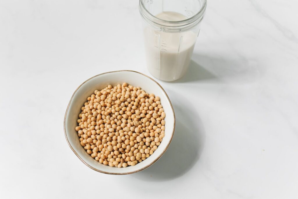 A bowl of dried soybeans beside a glass of soy milk on a pale marble surface